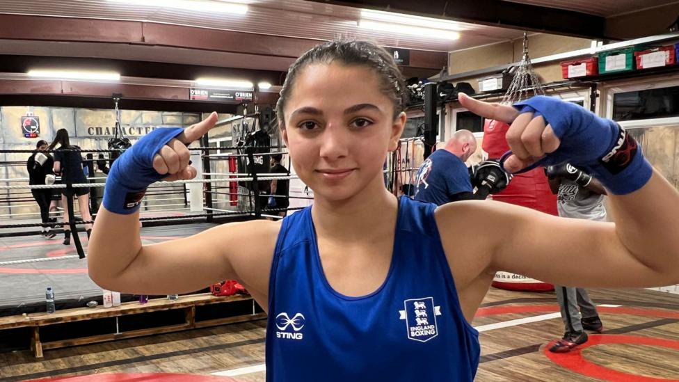 A girl wearing a blue vest stands outside a boxing ring, with her arms in a fighting stance.
