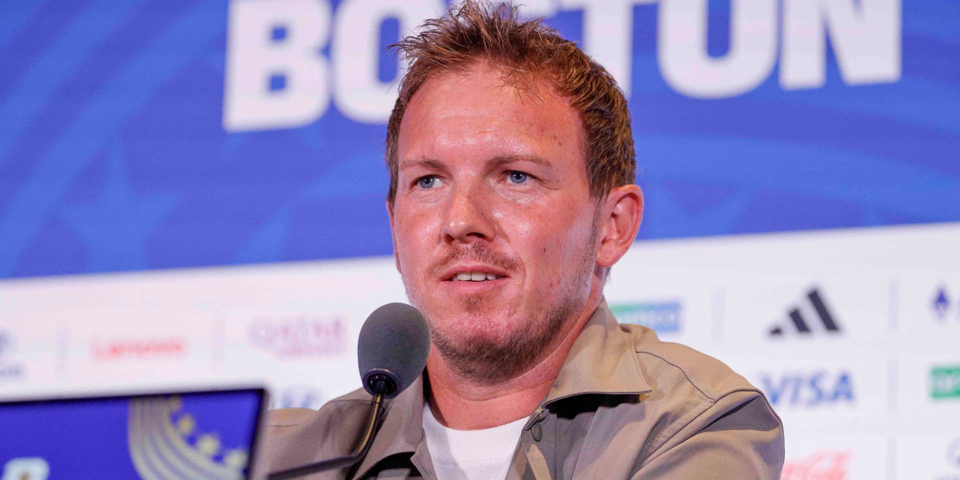 Julian Nagelsmann sitzt im hellbraunen Hemd vor Mikrofonen auf einer Pressekonferenz.