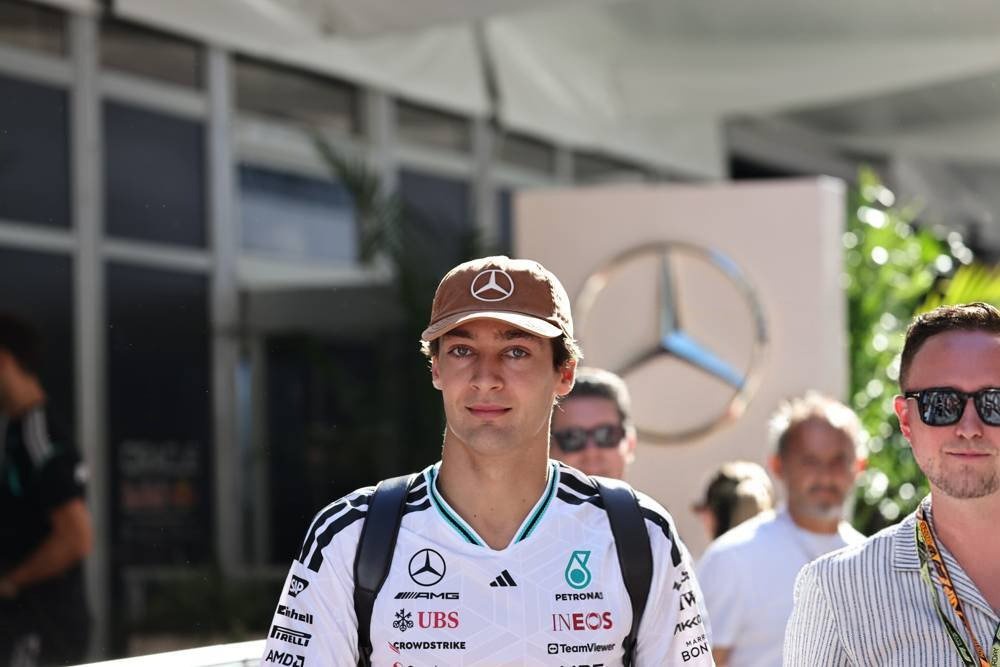 A man in a Mercedes-AMG Petronas F1 team cap and white race shirt with sponsor logos (UBS, INEOS, CrowdStrike) walks through a paddock area with a Mercedes s…