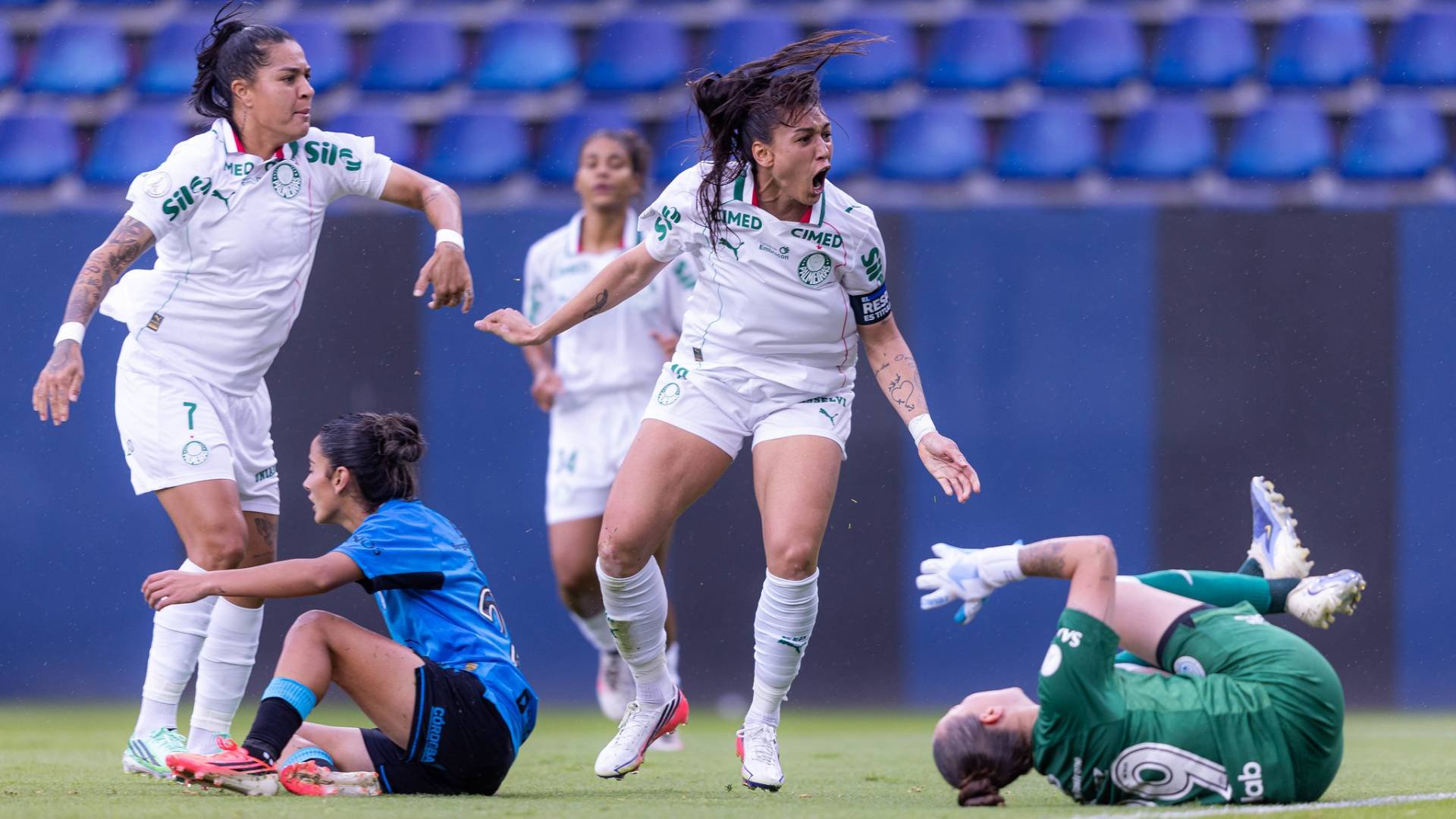 Jogadoras de futebol feminino em campo. Uma atleta de camisa branca e shorts brancos, com cabelo preso, grita com a boca aberta e braços flexionados. Outra jogadora de camisa branca e shorts brancos, com cabelo preso, observa. Uma terceira, de camisa azul e shorts pretos, está sentada no chão. Uma goleira de camisa verde e luvas brancas está deitada de costas no gramado. Cadeiras azuis vazias ao fundo