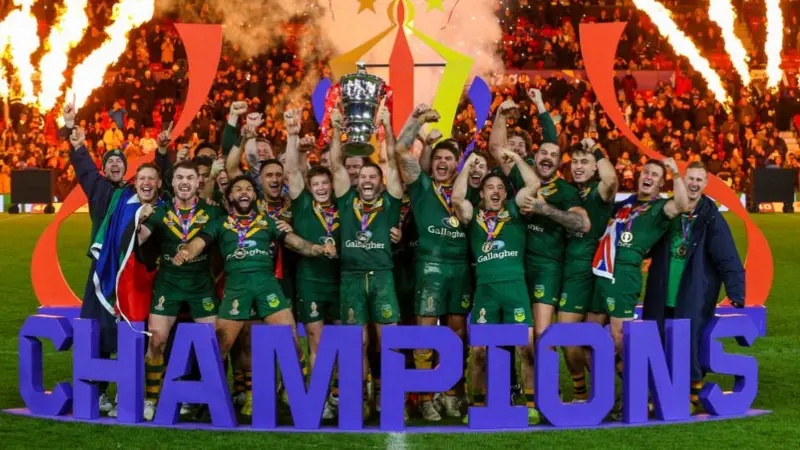 Australia's players, wearing green kits, lift the silver World Cup trophy behind the word champions, written in blue capital letters