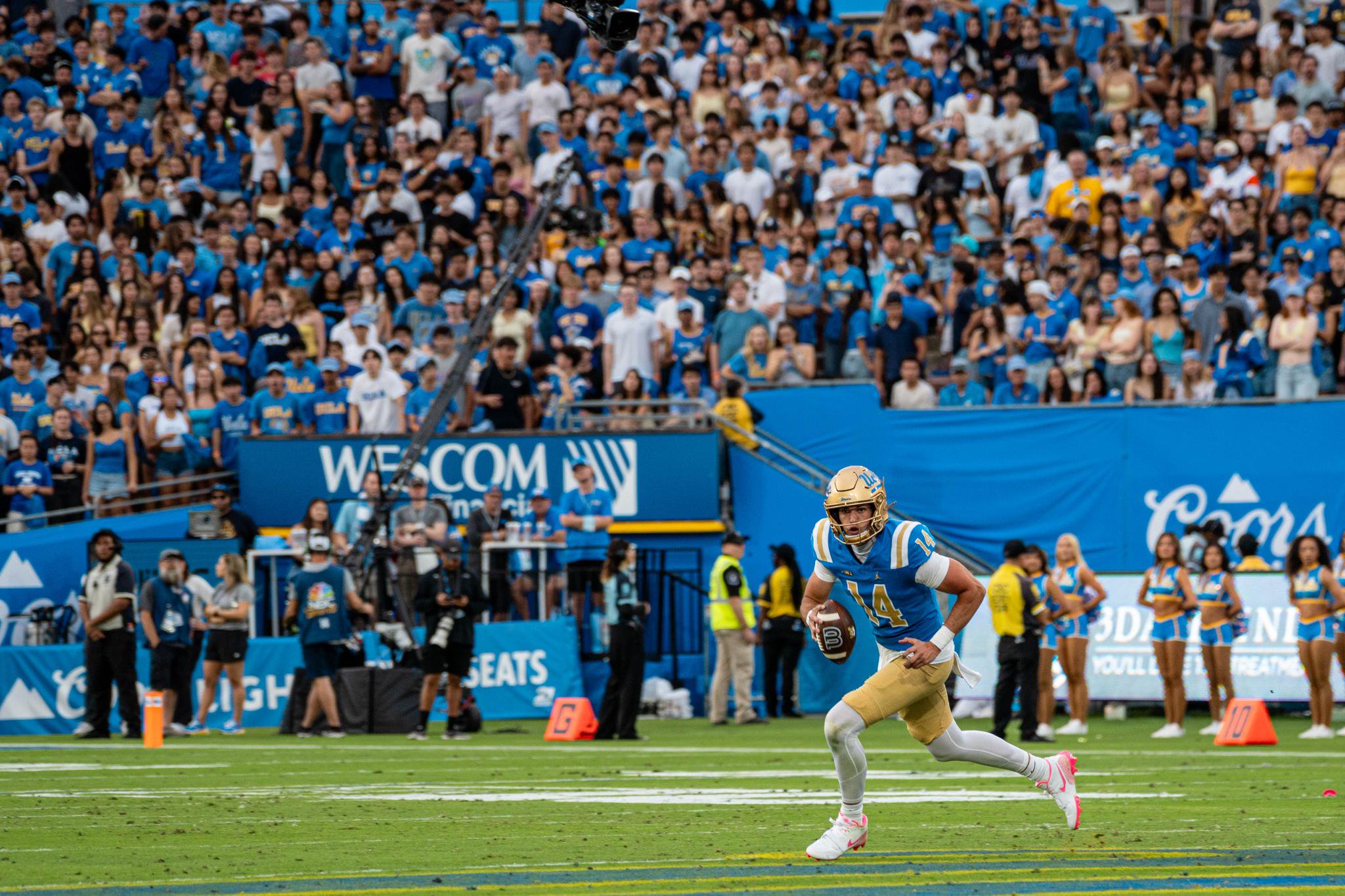 A UCLA quarterback in a blue and gold uniform running with the football during a game, with a large crowd in the background.