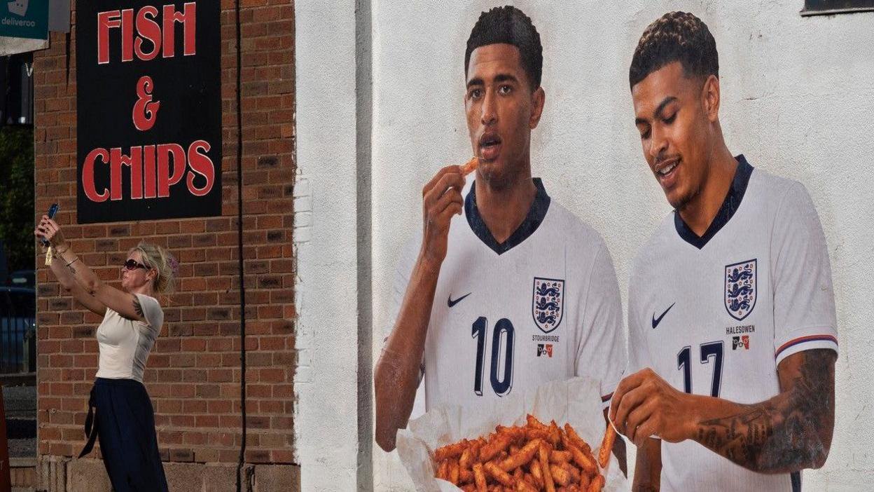 Jude Bellingham and Morgan Rogers on the side of a fish and chip shop in Birmingham. They are tucking into some chips while a woman takes a selfie with them.
