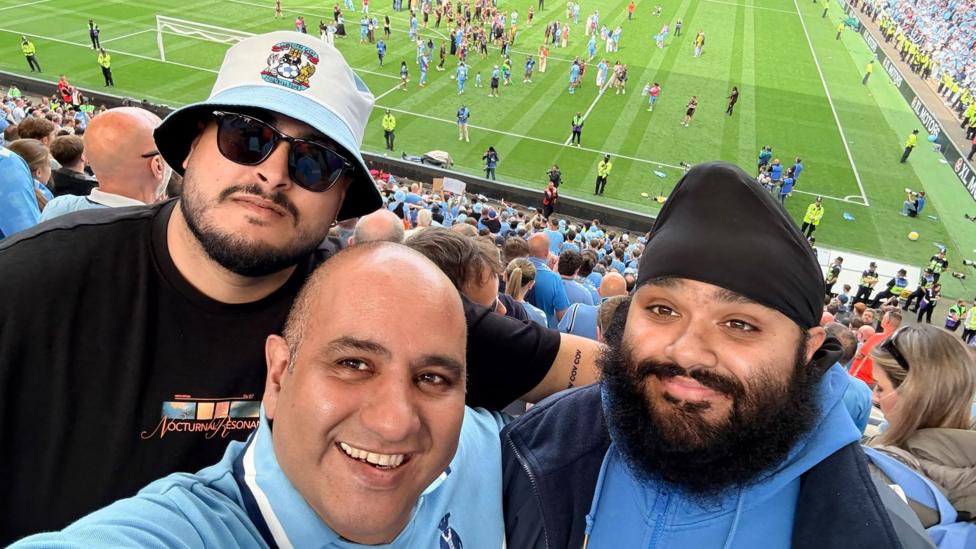 Three men taking a selfie in front of a football pitch. On the left, the man has black facial hair and is wearing ark sunglasses and a light blue and white bucket hat. To his right is a bald man wearing a light blue t-shirt. 