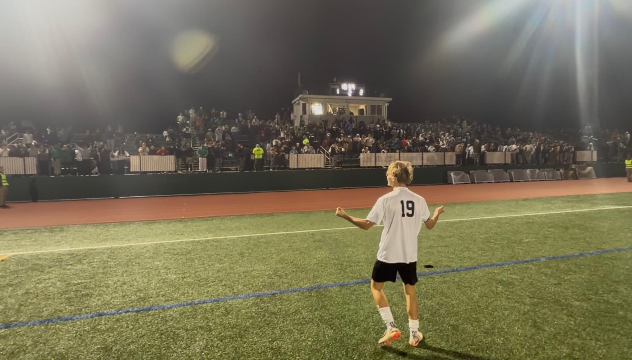 A soccer player wearing jersey number 19 celebrates a victory on the field at night, facing a crowded stadium with bright floodlights overhead.