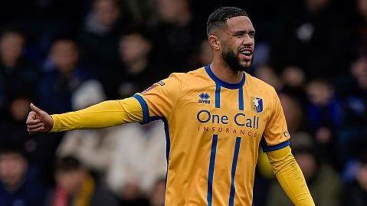 Tyler Roberts in action for Mansfield Town. He is giving a thumbs-up to a team-mate who is out of the picture. He is wearing a yellow shirt with two blue stripes down the middle and blue shorts. 