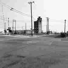 Black-and-white photo of an urban street with intersecting power lines, traffic lights, and a tall, narrow, derelict building in the center. Nearby, there are industrial structures and a car parked on the right side of the road. The overall scene looks deserted.
