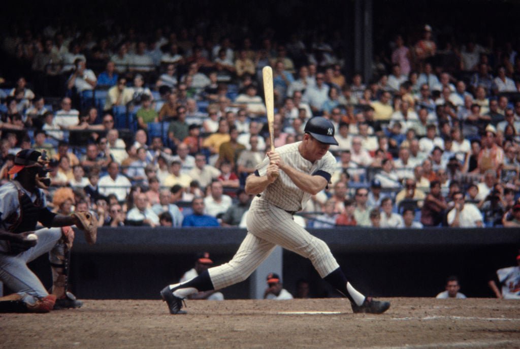 A baseball player in a striped uniform swings the bat during a game. A catcher and umpire are positioned behind him. The stands are filled with spectators watching the action.