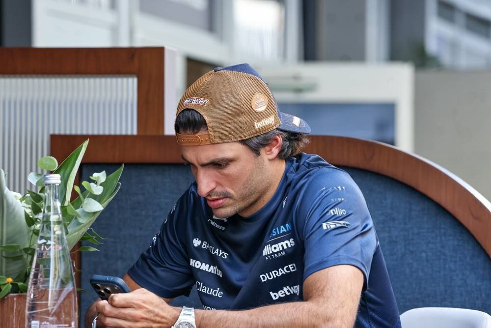 A Williams F1 driver in a navy team shirt and tan Betway cap, looking at his phone at a paddock table