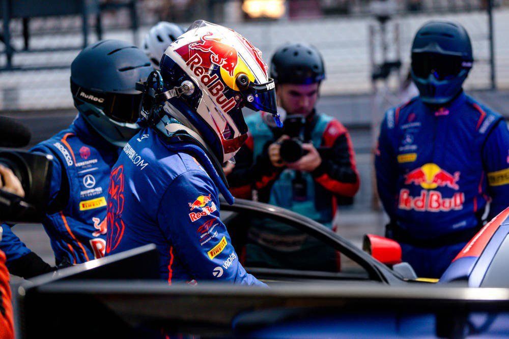 Driver in Red Bull racing suit and helmet beside a car in the pit lane, with crew members nearby; Verstappen-related 24-hour race coverage