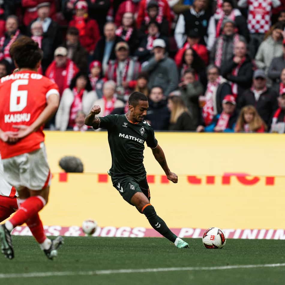 Arthur em ação durante partida de futebol, chutando a bola com torcedores vestindo vermelho e branco ao fundo.