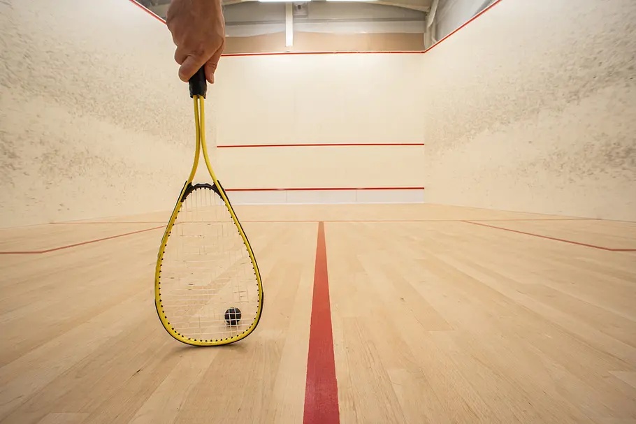 Eine Hand hält einen gelben Squashschläger vor einem schwarzen Ball auf dem Holzboden eines Courts mit roten Linien.