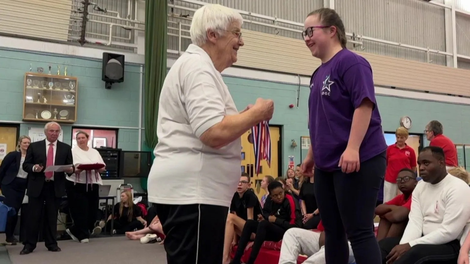 Kay Salter presents a medal to a female gymnast in front of other athletes and judges in a gym.